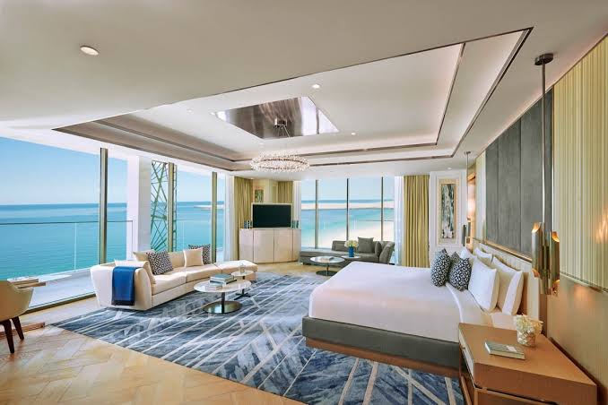 A contemporary hotel suite bedroom and living area featuring a large bed, a white sofa on a patterned blue rug, and massive windows offering a sweeping view of the ocean, all grouped under most prestigious hotel suites
