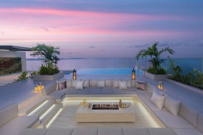A luxurious outdoor terrace seating area with a sunken fire pit surrounded by white cushioned sofas, modern lanterns, and a panoramic view of the calm ocean under a colorful sunset sky, all grouped under most prestigious hotel suites