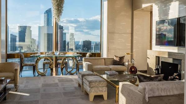 A sophisticated hotel living room with a glass-topped dining table featuring ornate golden bases, a cozy sofa, armchairs, a fireplace, and large windows framing a high-rise city skyline, all grouped under most prestigious hotel suites
