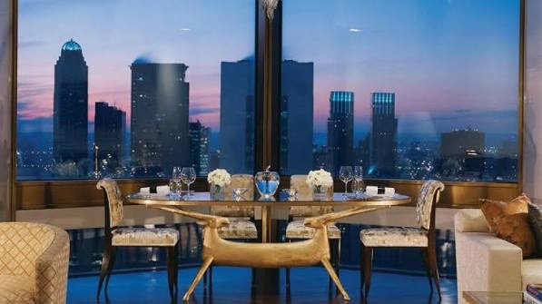 A luxurious dining table set with elegant chairs and glassware, positioned in front of large floor-to-ceiling windows overlooking a city skyline at dusk, all grouped under most prestigious hotel suites