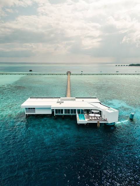 A modern white overwater bungalow structure built on stilts in the middle of a turquoise ocean, connected to a long wooden pier under a cloudy sky, all grouped under most prestigious hotel suites