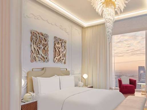 A bright and elegant bedroom featuring a plush white bed with a beige upholstered headboard, decorative abstract wooden wall panels, a cascading crystal chandelier, and a large window with sheer curtains showing a city skyline at sunset, all grouped under most prestigious hotel suites