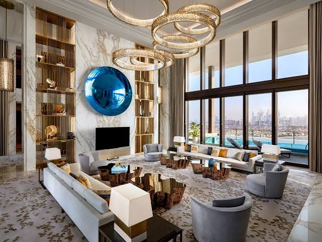 A grand, high-ceilinged hotel suite living room featuring white marble walls, a striking blue circular art piece, a large flat-screen television, unique metallic ring chandeliers, and floor-to-ceiling windows overlooking a city skyline and water, all grouped under most prestigious hotel suites