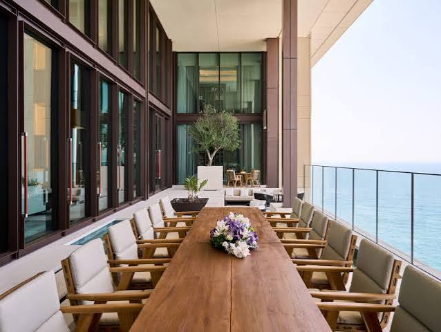 A long wooden outdoor dining table set with a floral centerpiece on a wide terrace flanked by comfortable armchairs, large glass doors, and a panoramic ocean view, all grouped under most prestigious hotel suites