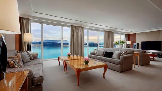 A spacious, elegant living room featuring comfortable gray sofas, two glass-topped wooden coffee tables with flower arrangements, and wide floor-to-ceiling windows overlooking a scenic waterfront and distant mountains, all grouped under most prestigious hotel suites