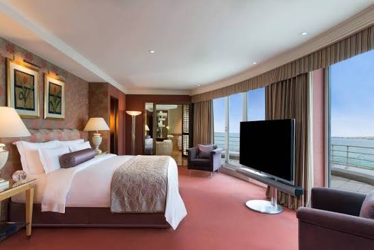 A luxurious hotel bedroom featuring a plush bed with patterned pillows, a large television facing the bed, framed artwork on a patterned wallpaper wall, and expansive floor-to-ceiling windows with sheer and heavy curtains offering an ocean view, all grouped under most prestigious hotel suites