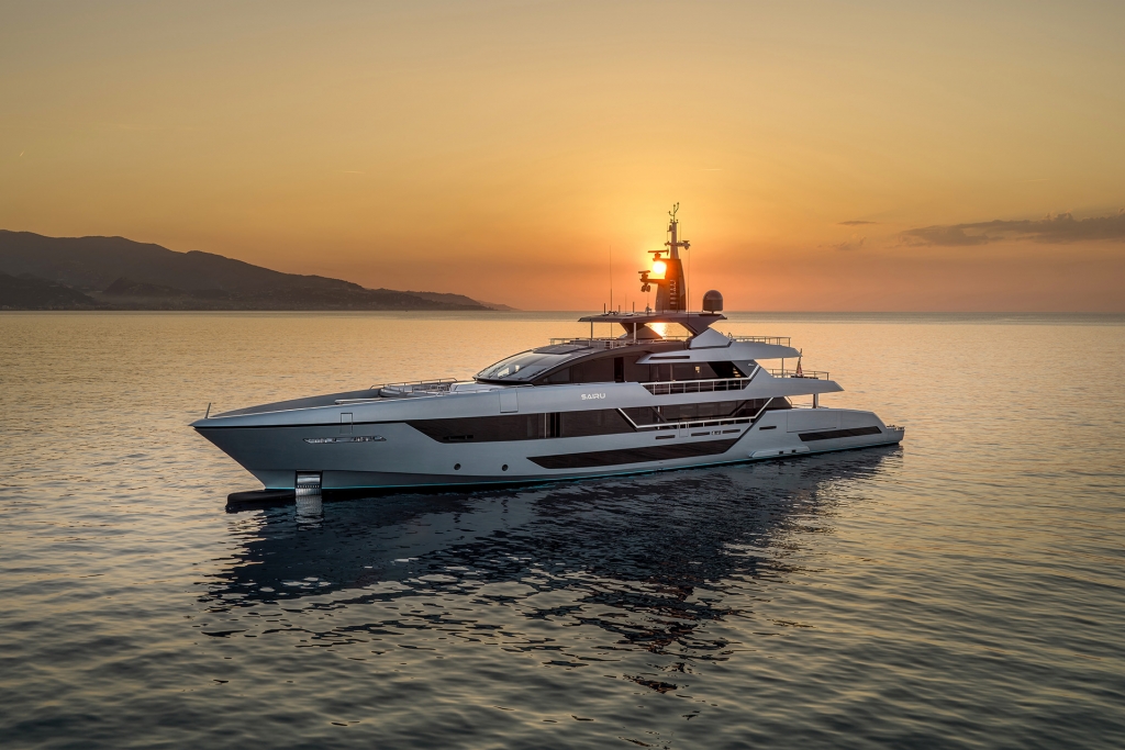 A long, sleek superyacht is anchored at sea during a dramatic golden-orange sunset, with silhouetted hills in the distance. monaco yacht show