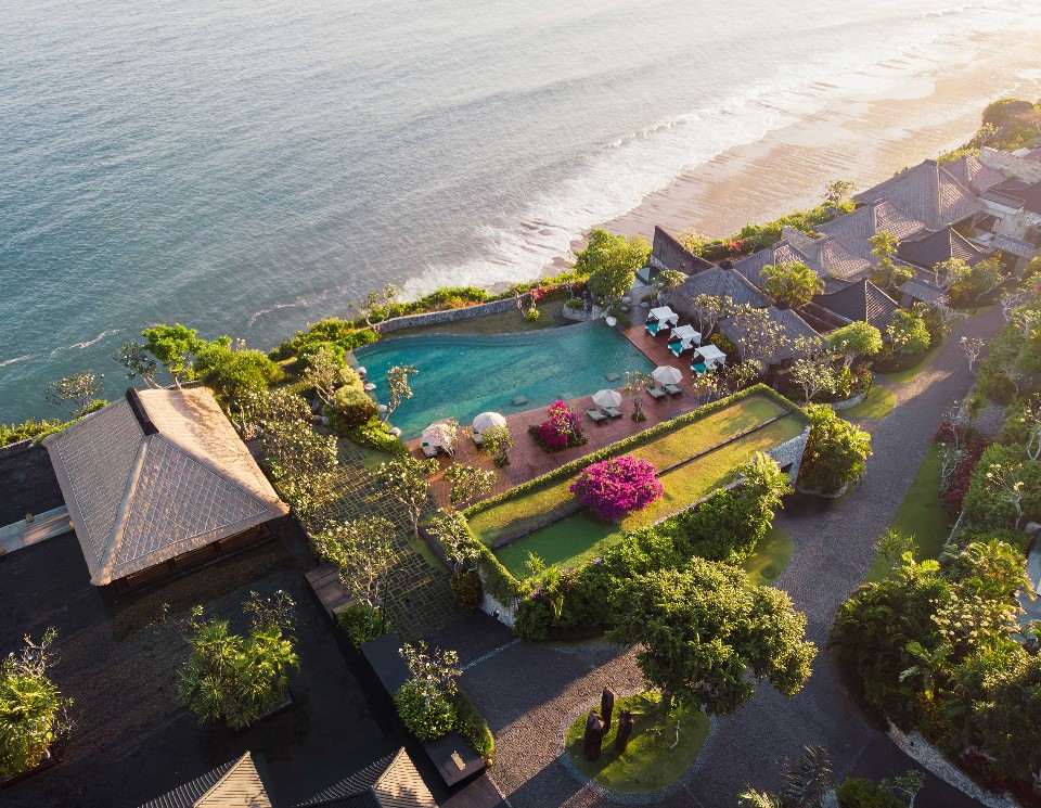 An elevated view of a tropical beachfront resort, featuring numerous thatched-roof villas, a lagoon-style swimming pool, and sun loungers set amidst palm trees and vibrant magenta bougainvillea, with gentle waves lapping the shore at sunrise. Luxury hotel chains.