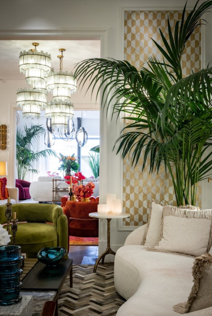 An elegant showroom display showcasing a curved cream sofa, a green velvet armchair, statement lighting, and a large palm tree against patterned wallpaper at luxury furniture showrooms in Monaco.