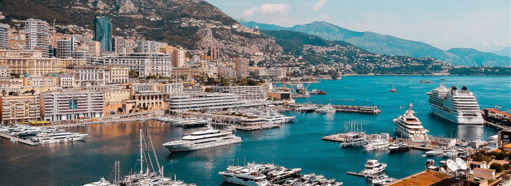 A wide, elevated coastal view of the Port of Monaco, filled with numerous superyachts, tenders, and a large cruise ship, with the city's buildings rising sharply up the steep, green mountains under a bright blue sky. monaco yacht show