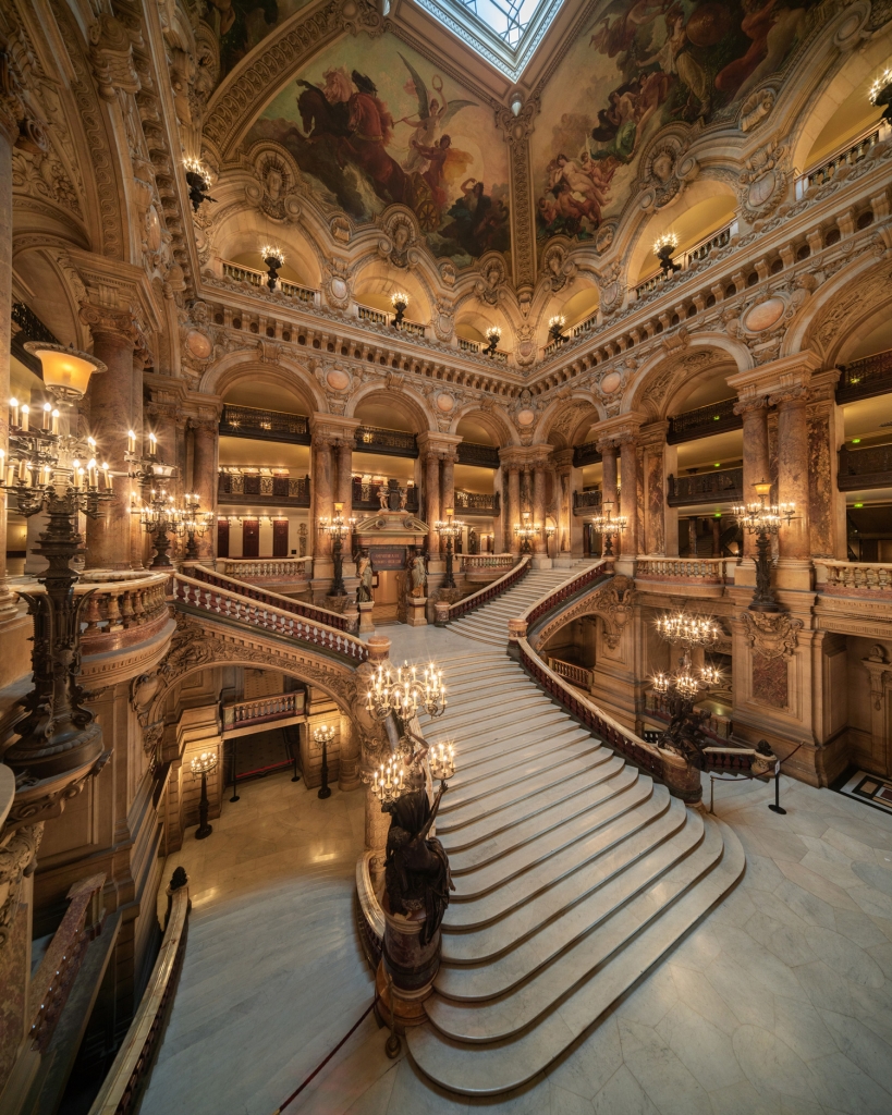 An opulent interior view of the grand staircase at the Opéra Garnier in Paris, showcasing marble stairs, elaborate ornate gold columns, detailed vaulted ceilings with classical frescoes, and glowing chandeliers. Paris design week.