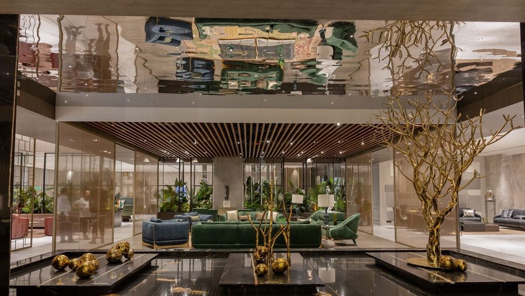 A luxurious showroom interior with a polished mirrored ceiling reflecting the space, featuring a dark green velvet sofa grouping, gold accent decor, and polished black marble display tables at luxury furniture showrooms in Hyderabad.