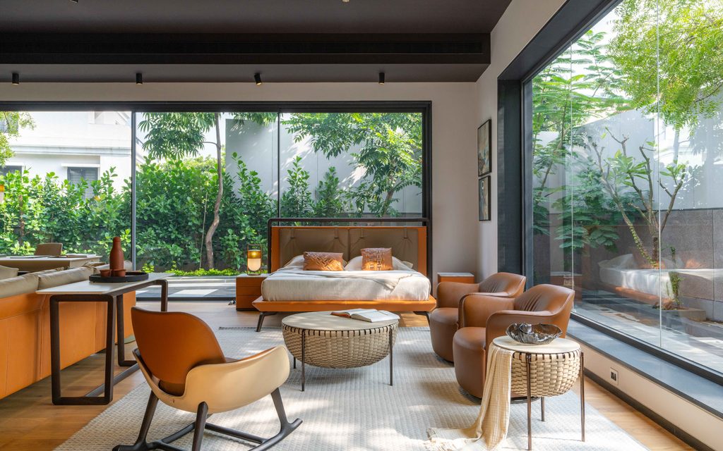 A bright, open-plan luxury bedroom and lounge area featuring a brown leather bed, matching armchairs, a woven coffee table, and floor-to-ceiling glass walls overlooking lush greenery at luxury furniture showrooms in Hyderabad.