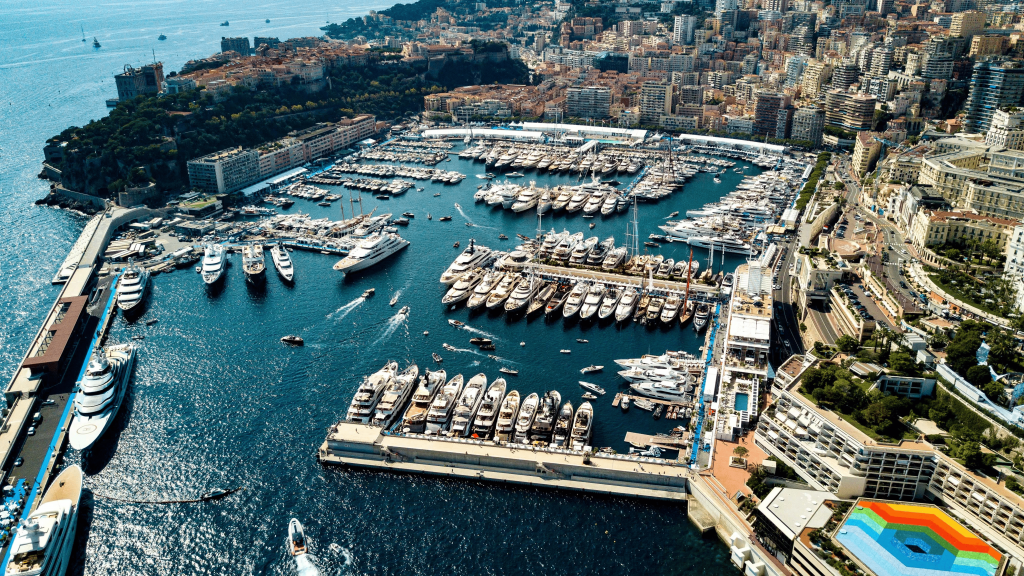 An elevated, panoramic aerial view of Port Hercules in Monaco, densely packed with hundreds of superyachts and smaller vessels, surrounded by the high-rise buildings of the city built into the hillside. monaco yacht show