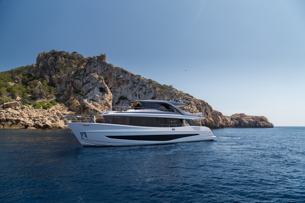 A medium-long shot of a modern white motor yacht cruising across deep blue water, viewed from its starboard quarter. The yacht has multiple decks, large windows, and a sleek profile. It is navigating along a rugged, rocky coastline with a historical structure visible on the cliff top under a clear sky. Cannes Yachting Festival.