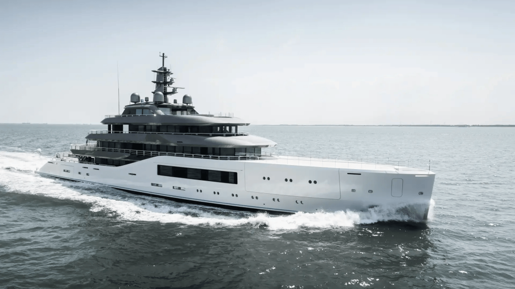 A side-profile view of a large, modern superyacht cruising at speed on the open ocean, with a white hull and a tiered, grey superstructure under a bright, hazy sky. The image has a slightly desaturated, monochromatic feel. monaco yacht show