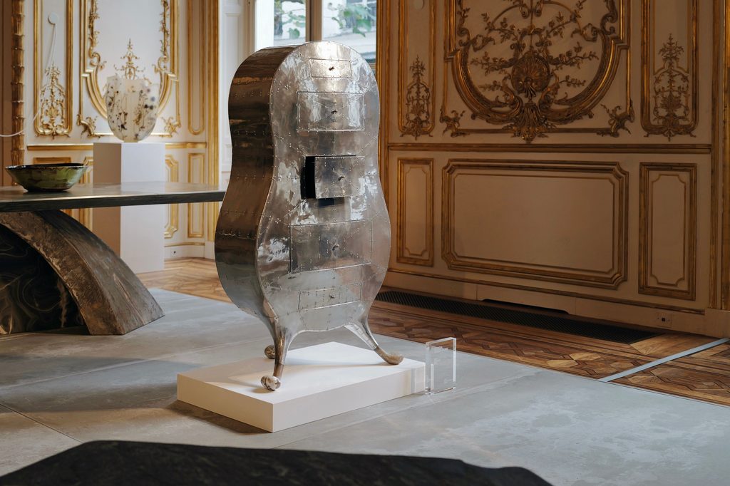 A metallic silver sculpted cabinet with drawers displayed on a plinth inside a room with gold-embellished walls and classical decor Design Miami.Paris 2026