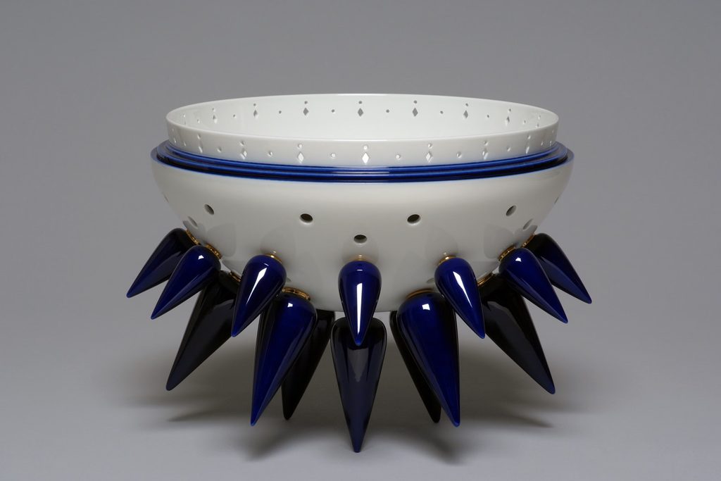 A decorative white ceramic bowl featuring gold accents and sharp, dark blue cone-shaped spikes around its lower rim Design Miami.Paris 2026