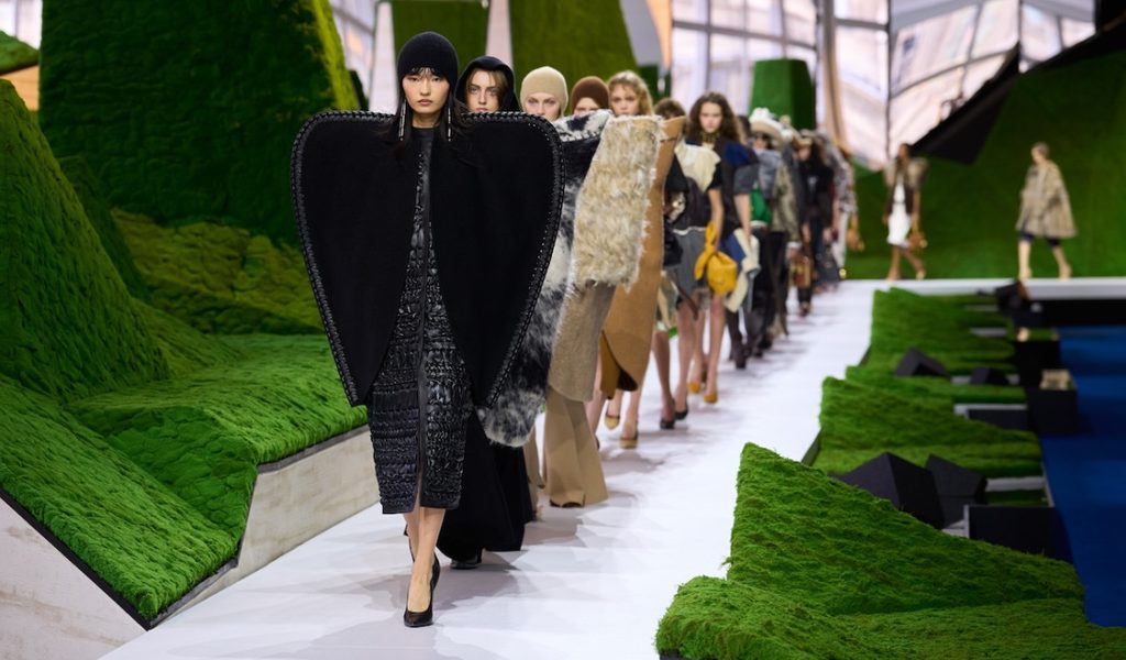 Models walking on a white runway flanked by green moss-covered hills, led by a model in a black structured cape and cap at Paris Fashion Week 2026