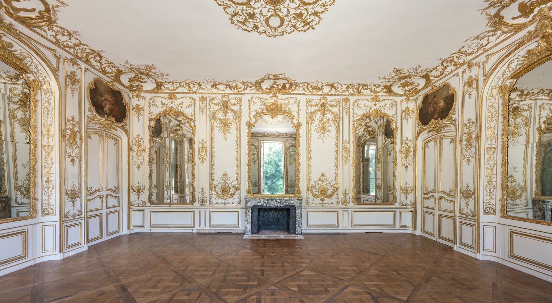 An opulent historical room with ornate gold leaf wall moldings, large gilded mirrors, a marble fireplace, and intricate parquet flooring Design Miami.Paris 2026.