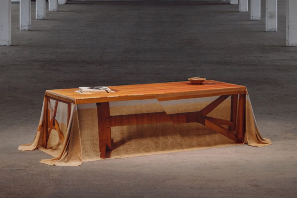 A wooden coffee table partially covered with a sheer, semi-transparent tablecloth printed with table leg graphics in a spacious industrial setting Design Miami.Paris 2026