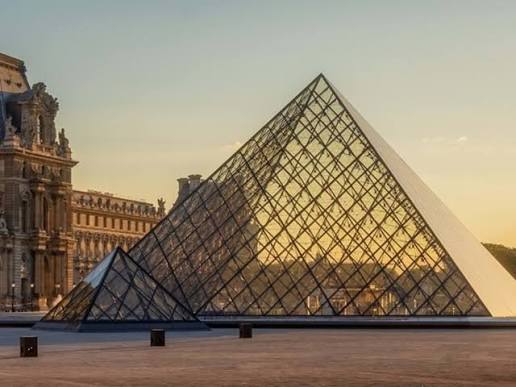 The iconic glass and metal pyramids of the Louvre Museum at sunrise, with surrounding historic stone architecture