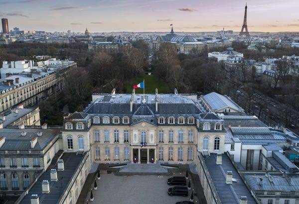 An aerial view of a grand classical French palace with a courtyard and the French flag flying on top, overlooking the cityscape of Paris with the Eiffel Tower in the distance.