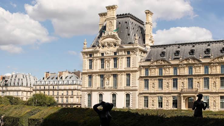A classic stone building with detailed architectural ornamentation, large windows, and a dark slate mansard roof under a bright cloudy sky.