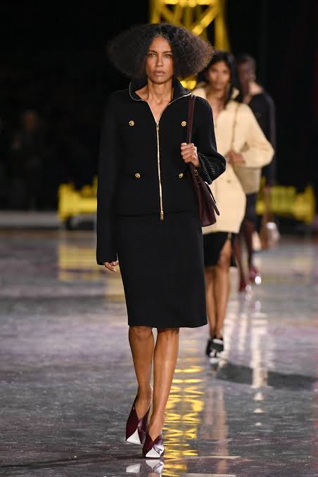 A model with voluminous dark hair wearing a black zippered jacket and matching knee-length skirt on a runway at Paris Fashion Week 2026