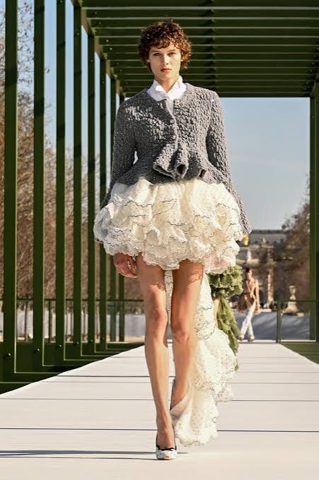 A model wearing a textured grey knit cardigan and a voluminous ruffled white miniskirt walking on an outdoor runway at Paris Fashion Week 2026