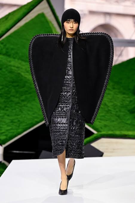 A model with a black beanie wearing a unique black structured cape with a quilted dress walking on a white runway at Paris Fashion Week 2026