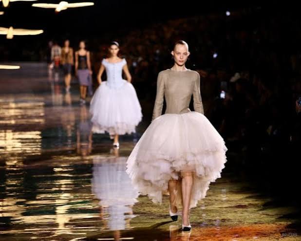 A model wearing a beige long-sleeved top and a layered, voluminous white tulle skirt walking along a reflective runway at Paris Fashion Week 2026
