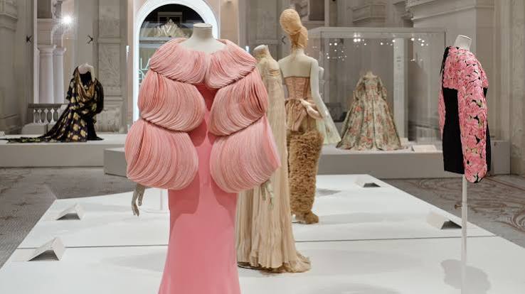 Mannequins displaying haute couture fashion items, including a pink dress with layered, voluminous rounded sleeves, inside an exhibition hall.