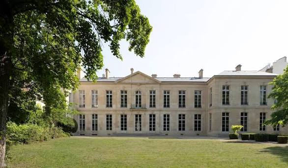 Exterior view of a grand French classical estate surrounded by lush green trees and a grassy lawn Design Miami.Paris 2026