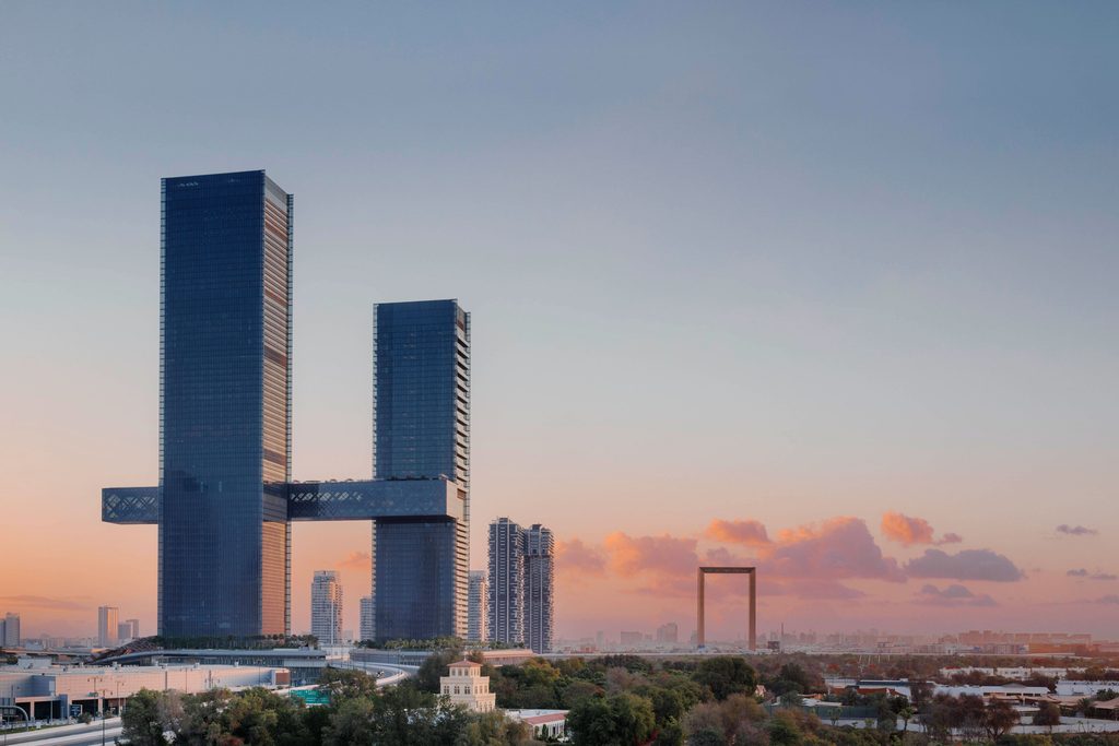Two modern skyscrapers connected by a large horizontal skybridge at sunset, with Dubai Frame in the background.