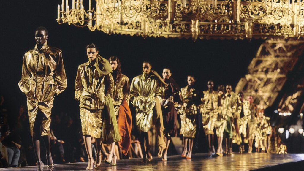 Models walking down a golden-lit runway wearing metallic gold outfits under a grand decorative structure at Paris Fashion Week 2026